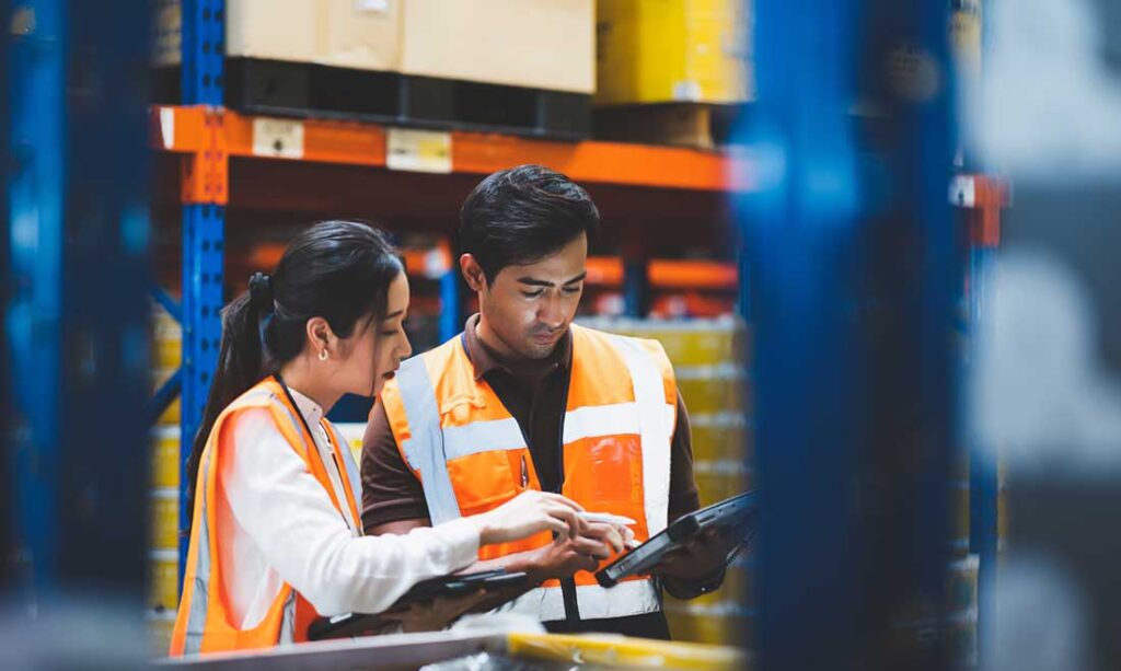 Warehouse employee and manager checking stock and inventory using digital tablet in retail warehouse full of shelves with goods.